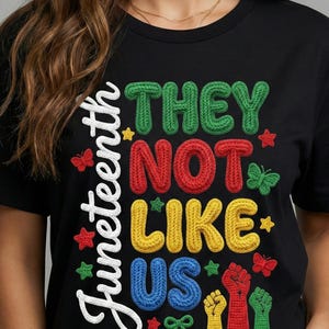May include: Black t-shirt with the text "Juneteenth" in white script and the phrase "THEY NOT LIKE US" in colorful, textured letters. The design includes stars, butterflies, and raised fists.
