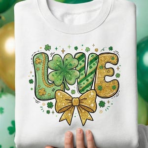 Puede incluir: Sudadera blanca con un diseño del Día de San Patricio. La palabra "LOVE" está escrita en purpurina verde y dorada, con un trébol de cuatro hojas que reemplaza la "O". Un lazo dorado en la parte inferior, con tréboles y estrellas verdes.