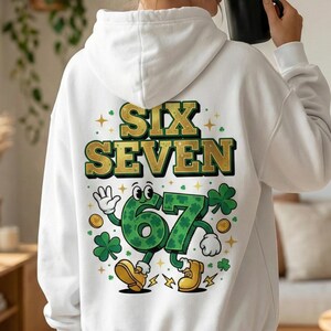 May include: White hoodie with the words "SIX SEVEN" in gold lettering. The number "67" is depicted as a cartoon character with a green shamrock design, wearing yellow shoes, and surrounded by shamrocks and gold coins.