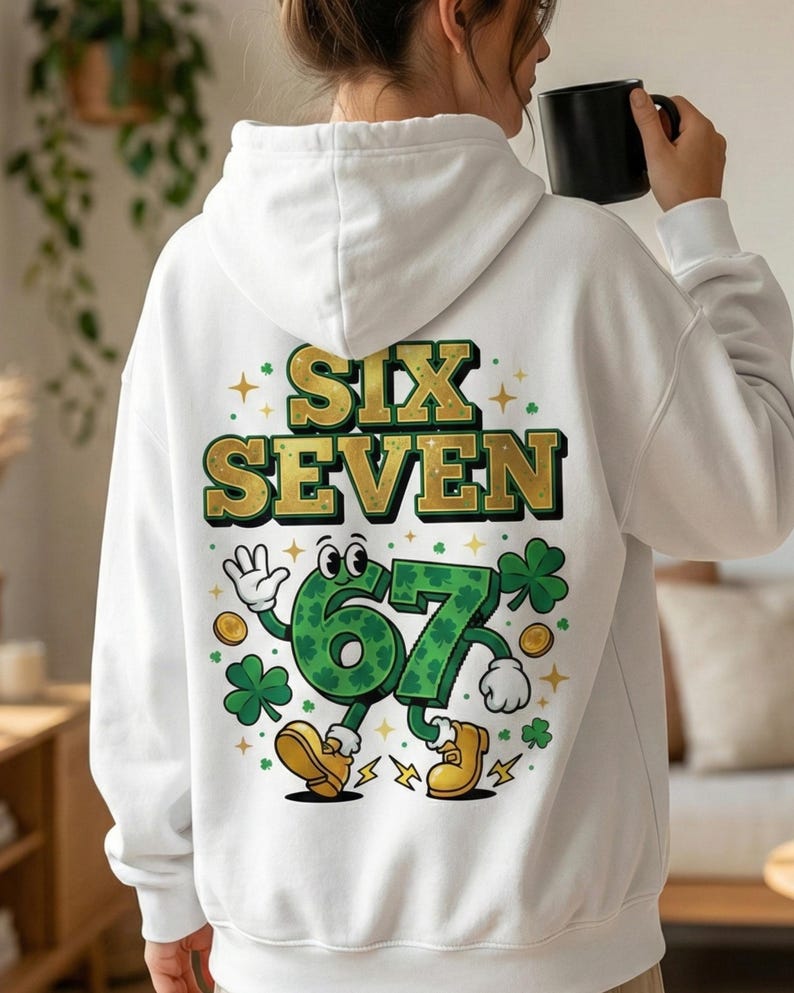 May include: White hoodie with the words "SIX SEVEN" in gold lettering. The number "67" is depicted as a cartoon character with a green shamrock design, wearing yellow shoes, and surrounded by shamrocks and gold coins.