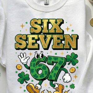 May include: White sweatshirt with a St. Patrick's Day design. The design features the words "SIX SEVEN" in gold and green, with a cartoon "67" wearing shoes and waving. Includes shamrocks, gold coins, and stars.