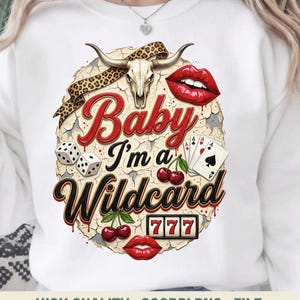 Op de afbeelding: Witte sweatshirt met een grafisch ontwerp met een schedel van een stier, rode lippen, dobbelstenen, speelkaarten, kersen en de tekst "Baby I'm a Wildcard". Het ontwerp heeft een vintage uitstraling.