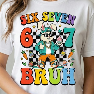 以下が含まれることがあります： 「SIX SEVEN BRUH」の文字とサングラス、帽子、金のネックレスをつけた漫画のウサギが描かれた、カラフルなグラフィックの白いTシャツ。ウサギはカップケーキを持っています。ニンジンと卵を使ったイースターをテーマにしたデザイン。