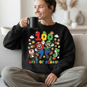 May include: Black sweatshirt featuring a colorful graphic of Mario, Luigi, and Yoshi with the text "100 DAYS OF SCHOOL". The design includes stars, mushrooms, and coins. The sweatshirt is worn by a person.