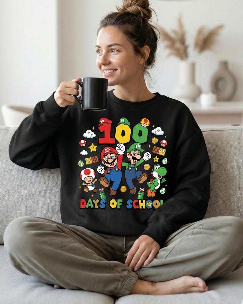 May include: Black sweatshirt featuring a colorful graphic of Mario, Luigi, and Yoshi with the text "100 DAYS OF SCHOOL". The design includes stars, mushrooms, and coins. The sweatshirt is worn by a person.