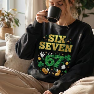 May include: Black sweatshirt with the text "SIX SEVEN" in gold and green. The number "67" is designed with a cartoon-like appearance, featuring hands, shoes, and shamrocks. The sweatshirt is worn by a person.