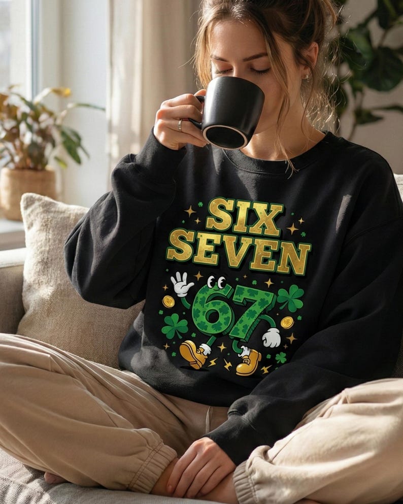 May include: Black sweatshirt with the text "SIX SEVEN" in gold and green. The number "67" is designed with a cartoon-like appearance, featuring hands, shoes, and shamrocks. The sweatshirt is worn by a person.