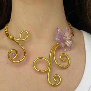 Könnte beinhalten: Ein goldfarbenes Draht-Collier mit floralem Akzent. Die Halskette hat ein wirbelndes Design mit einer lavendelfarbenen Blume und kleinen rosa Perlen. Der Schmuck wird von einer Person getragen, die ein weißes Tanktop trägt.