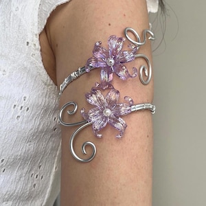 May include: A silver arm cuff with two lavender flower accents. The flowers have a pearl center and are attached to a silver wire design. A matching silver necklace with a flower accent is also visible.
