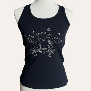 Könnte beinhalten: Schwarzes geripptes Tanktop mit einem Strass-Engel-Motiv und dem Wort "Angel". Das Top hat einen Rundhalsausschnitt und wird auf einer Schaufensterpuppe präsentiert. Mehrere Strasssterne befinden sich über dem Engel-Motiv.