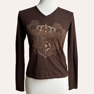 Könnte beinhalten: Ein braunes Langarm-V-Ausschnitt-Shirt mit einem verzierten Wappen-Design auf der Vorderseite. Das Wappen zeigt eine Krone, florale Elemente und funkelnde Verzierungen. Das Shirt wird auf einer Schaufensterpuppe präsentiert.