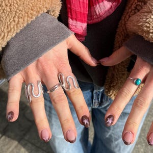 Könnte beinhalten: Nahaufnahme von Händen mit silbernen Ringen, darunter ein Wellendesign und ein Ring mit einem Edelstein. Die Nägel sind mit einem braunen French-Maniküre und Sternen verziert. Die Person trägt eine braune Jacke und blaue Jeans.