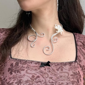Pode incluir: Um colar prateado com um design floral e uma pulseira de braço a condizer. O colar apresenta um design delicado e rodopiante com um acento floral. A pulseira de braço tem um design semelhante com pequenos acentos redondos. A modelo usa um top rosa floral.