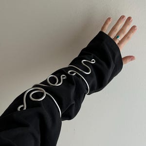 May include: Silver-toned arm cuffs with a swirling, abstract design are worn over a black sleeve. The cuffs have a polished finish and are arranged in a decorative pattern. A turquoise ring is visible on the hand.