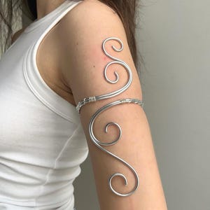 Könnte beinhalten: Ein silberner Armreif mit einem Wirbeldesign. Der Armreif umschließt den Oberarm und weist dekorative Locken auf. Der Schmuck besteht aus einem glänzenden, metallischen Material.