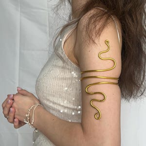 Könnte beinhalten: Goldfarbene Armspange mit einem wirbelnden, schlangenartigen Design. Die Spange umschließt den Oberarm mit einem einfachen Band und einem dekorativen, gebogenen Drahtelement. Der Schmuck wird am Arm getragen.