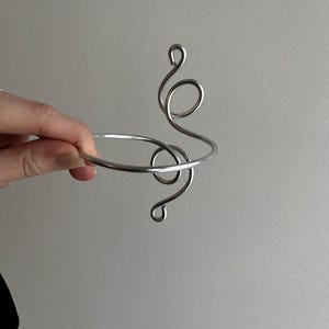 May include: A silver-colored metal bracelet with a decorative, swirling design. The bracelet has a circular band and an abstract, flowing pattern that extends upwards. The design includes several loops and curves, creating a unique and artistic look.