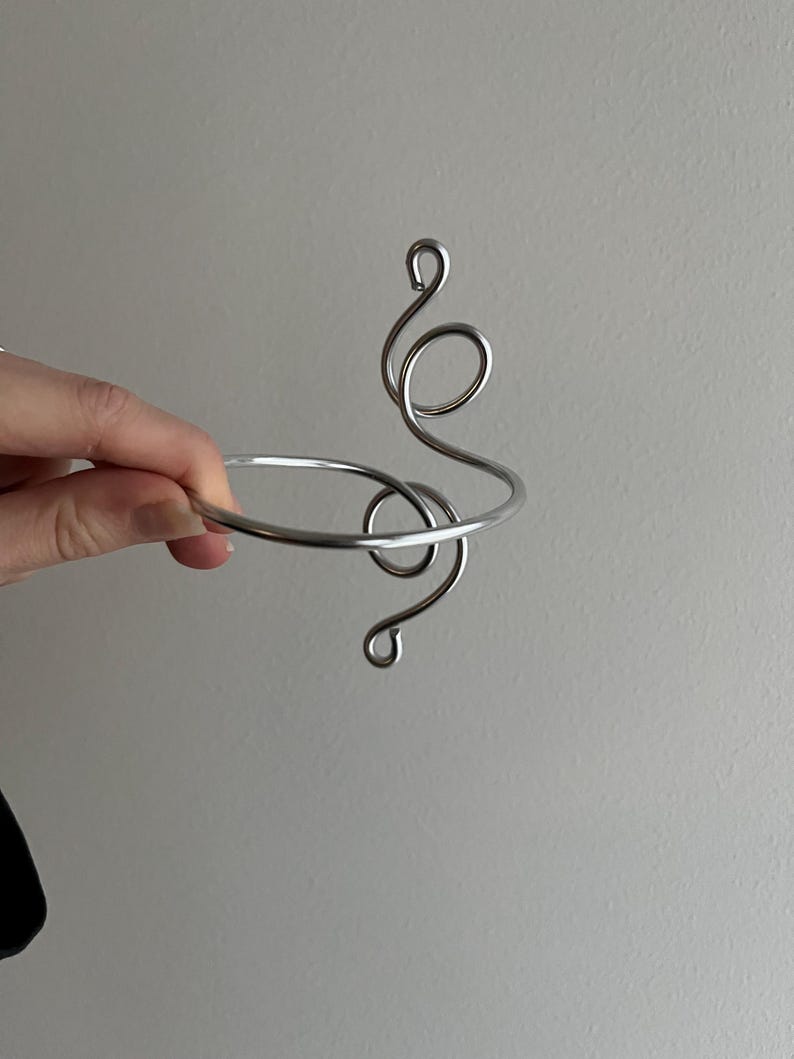 May include: A silver-colored metal bracelet with a decorative, swirling design. The bracelet has a circular band and an abstract, flowing pattern that extends upwards. The design includes several loops and curves, creating a unique and artistic look.