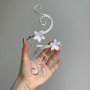 May include: A silver-toned arm cuff with a floral design. The cuff features two white flower accents and a dangling cluster of three small, white beads. The design includes swirling, decorative metalwork.