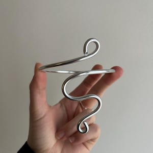 May include: A silver-toned arm cuff with a flowing, abstract design. The cuff features a curved, open design with a spiral and loop detail. The jewelry is held in a hand against a neutral background.