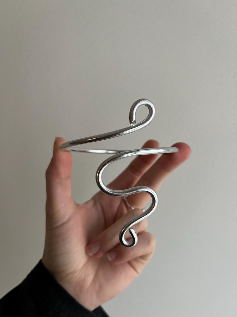May include: A silver-toned arm cuff with a flowing, abstract design. The cuff features a curved, open design with a spiral and loop detail. The jewelry is held in a hand against a neutral background.