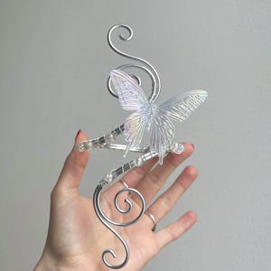 May include: A silver wire headpiece with a clear, iridescent butterfly and crystal embellishments. The headpiece has decorative swirls and is designed to be worn on the head. The butterfly has detailed wing patterns.