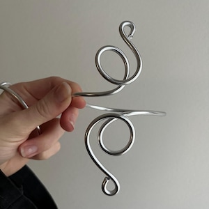 May include: A silver-toned arm cuff with a swirling, open design. The cuff features two large, rounded loops connected by a straight bar. The metal has a polished, reflective surface, creating a modern and elegant aesthetic.