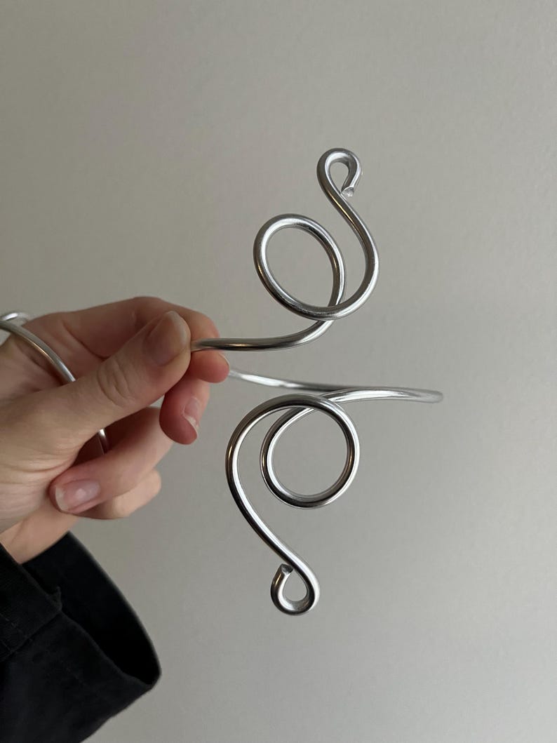 May include: A silver-toned arm cuff with a swirling, open design. The cuff features two large, rounded loops connected by a straight bar. The metal has a polished, reflective surface, creating a modern and elegant aesthetic.