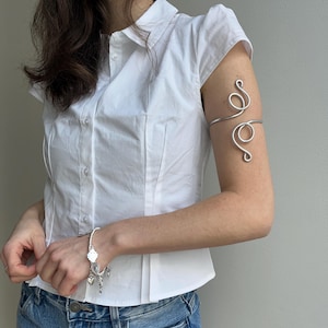 May include: A silver arm cuff with a swirling design and a silver bracelet with charms. The arm cuff is worn on the upper arm. The bracelet has heart and cross charms. The person is wearing a white button-down shirt and blue denim shorts.