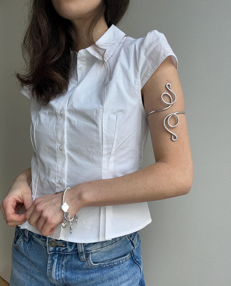 May include: A silver arm cuff with a swirling design and a silver bracelet with charms. The arm cuff is worn on the upper arm. The bracelet has heart and cross charms. The person is wearing a white button-down shirt and blue denim shorts.
