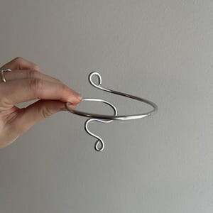May include: A silver-colored arm cuff with a minimalist design. The cuff features a curved, open design with a spiral detail at the top and a looping swirl at the bottom. The jewelry is held against a neutral background.