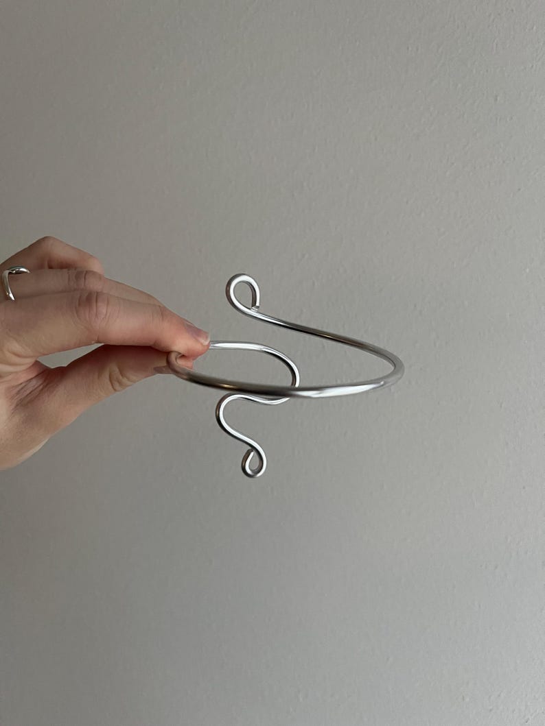 May include: A silver-colored arm cuff with a minimalist design. The cuff features a curved, open design with a spiral detail at the top and a looping swirl at the bottom. The jewelry is held against a neutral background.