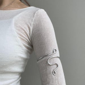 May include: A silver-colored arm cuff with a flowing, abstract design. The cuff wraps around the upper arm, featuring a curved line that forms a series of loops and swirls. The jewelry is worn on a light-colored long-sleeved shirt.