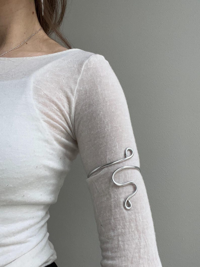 May include: A silver-colored arm cuff with a flowing, abstract design. The cuff wraps around the upper arm, featuring a curved line that forms a series of loops and swirls. The jewelry is worn on a light-colored long-sleeved shirt.