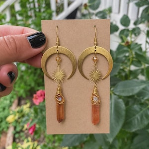Op de afbeelding: Een paar goudkleurige bungelende oorbellen met een halve maan bedel, een kleine zon bedel en een amberkleurig kristal omwikkeld met goudkleurig draad. De oorbellen hangen aan goudkleurige haakjes op een bruine kartonnen kaart.