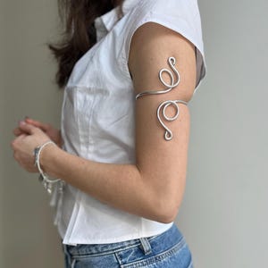 May include: Silver arm cuff with a swirling design. The cuff is worn on the upper arm and features a continuous, flowing pattern. The jewelry is made of a shiny, metallic material and is a decorative accessory.