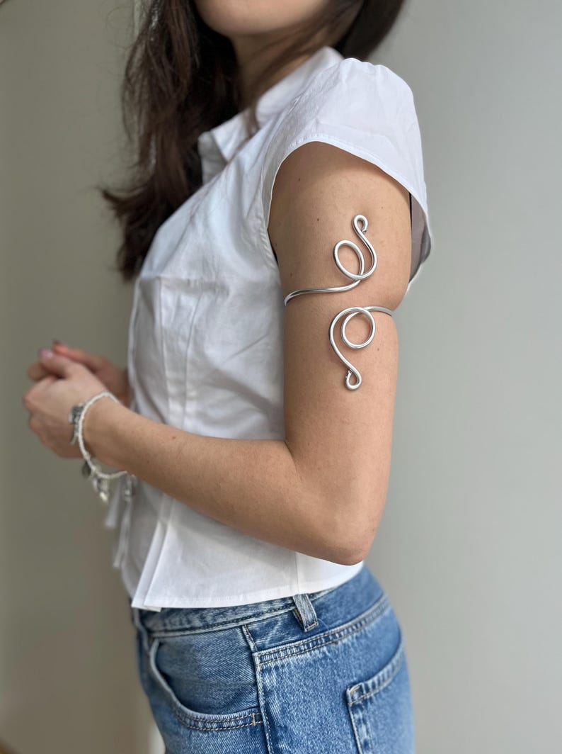 May include: Silver arm cuff with a swirling design. The cuff is worn on the upper arm and features a continuous, flowing pattern. The jewelry is made of a shiny, metallic material and is a decorative accessory.