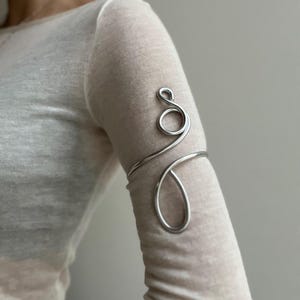 Op de afbeelding: Een zilverkleurige armmanchet met een wervelend ontwerp. De manchet wikkelt zich om de bovenarm en heeft een lus en een druppelvorm. De sieraden zijn gemaakt van een glanzend, metalen materiaal.