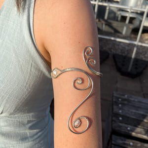 Könnte beinhalten: Silberfarbene Armspange mit filigranem, wirbelndem Design. Die Spange hat kleine, runde, perlenartige Akzente. Der Schmuck wird am Oberarm getragen und zeigt seinen eleganten, künstlerischen Stil. Das Design integriert anmutige Kurven und eine minimalistische Ästhetik.
