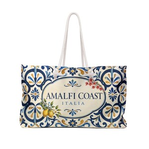 May include: A rectangular tote bag with white rope handles featuring a cream-colored center oval with the text AMALFI COAST ITALIA in dark blue. The bag is decorated with a blue, yellow, and white floral tile pattern and lemon illustrations.