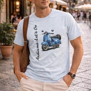 Könnte beinhalten: Hellblaues T-Shirt mit einer blauen Roller-Illustration und dem Text "Andiamo-Let's Go". Das Shirt besteht aus einem weichen Material und eignet sich für Freizeitkleidung. Das Design ist im Vintage-Stil.