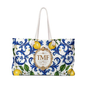 Può includere: Borsa tote bianca con un motivo floreale blu e bianco, limoni gialli e le iniziali "TMF" all'interno di una cornice dorata. La borsa ha manici in corda bianca e la scritta "EST. 2026".