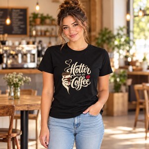 May include: A black short-sleeved t-shirt featuring a graphic print of a hand holding a coffee cup with steam rising in a heart shape. The text reads Hotter than my Coffee in a stylized cream font, accompanied by a small red lipstick kiss mark.