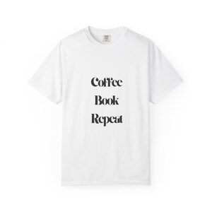 May include: A white t-shirt featuring the words "Coffee Book Repeat" in black, arranged vertically. The shirt has a crew neck and short sleeves. The text is in a bold, sans-serif font.