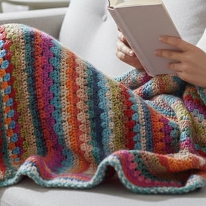 Pode incluir: Uma manta de crochê colorida com um padrão listrado em tons de azul, laranja, rosa e verde. A manta está sobre o colo de uma pessoa, com um livro nas mãos. A borda da manta tem um design ondulado.