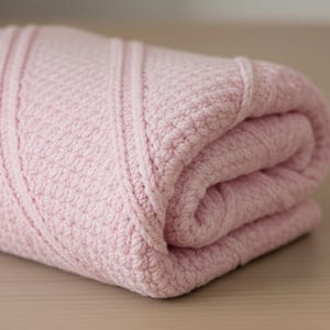 May include: A soft, light pink knitted blanket featuring a textured pattern of diagonal stitches interspersed with smooth, vertical ribbed lines. The fabric is folded over itself, showcasing the consistent yarn weave and the pastel color throughout.