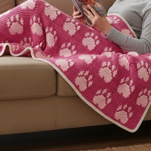 Op de afbeelding: Een roze gehaakte deken met grote witte pootafdrukken. De deken heeft een witte rand en is gedrapeerd over een persoon die op een bruine bank zit. Een persoon leest een boek.