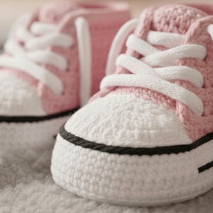 Może przedstawiać: Zbliżenie na dwa ręcznie robione buciki dla niemowląt. Buty są szydełkowane w różowo-białej kolorystyce, z białymi sznurowadłami i czarnym paskiem wzdłuż podeszwy. Trampeczki są zaprojektowane tak, aby przypominały klasyczne płócienne buty.