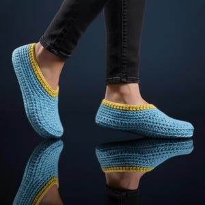 May include: Light blue crochet slippers with yellow edging. The image shows the slippers on a person's feet, reflected in a dark, glossy surface. The slippers are likely handmade, offering a cosy, comfortable design.