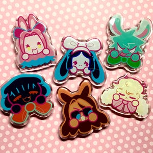 May include: A collection of six colorful acrylic pins featuring cartoon characters with bunny ears and bows. The pins showcase a variety of colors, including pink, blue, green, and brown, with detailed facial expressions and outlines. The background is a pink polka dot pattern.